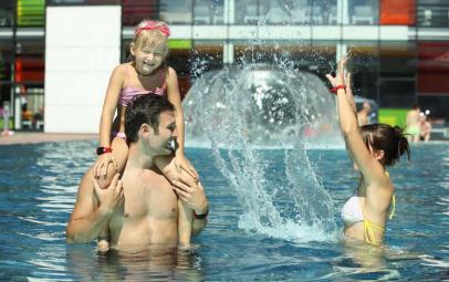 Ein Mann trägt ein lächelndes junges Mädchen auf seinen Schultern in einem Pool, während eine Frau in der Nähe Wasser spritzt.