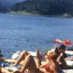 Menschen, die sich auf einer Wiese am Ufer eines Sees sonnen und entspannen, mit Bäumen und Hügeln im Hintergrund.