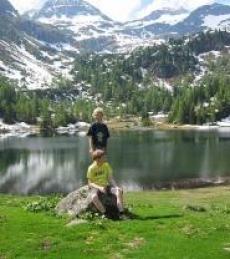 Zwei Kinder befinden sich im Freien an einem See, wobei eines auf einem Felsen sitzt und das andere dahinter steht, umgeben von Gras, Bäumen und schneebedeckten Bergen im Hintergrund.