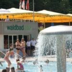 Menschen schwimmen und entspannen sich in einem Freibad mit einer Wasserfontäne und einem Gebäude mit der Aufschrift "Waldbad Enz" im Hintergrund. Gelbe Vordächer spenden Schatten.