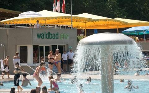 Menschen schwimmen und entspannen sich in einem Freibad mit einer Wasserfontäne und einem Gebäude mit der Aufschrift "Waldbad Enz" im Hintergrund. Gelbe Vordächer spenden Schatten.