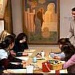 Eine Gruppe von Menschen sitzt an einem Tisch und arbeitet an Kunstprojekten, während ein Mann mit einer Tasse in der Hand in einem mit Gemälden dekorierten Raum steht.