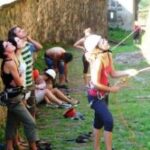 Mehrere Personen in Kletterausrüstung stehen und sitzen an einer Steinwand, einige schauen nach oben, während eine Person im Vordergrund ein Kletterseil hält.