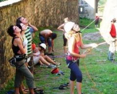 Mehrere Personen in Kletterausrüstung stehen und sitzen an einer Steinwand, einige schauen nach oben, während eine Person im Vordergrund ein Kletterseil hält.