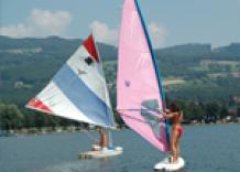 Zwei Personen beim Windsurfen auf einem See, jede auf einem eigenen Brett mit bunten Segeln, mit Hügeln und Bäumen im Hintergrund.