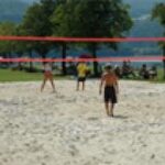 Menschen spielen Beachvolleyball auf einem Sandplatz im Freien, mit Bäumen, Gras und einem Gewässer im Hintergrund.
