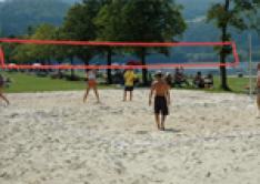 Menschen spielen Beachvolleyball auf einem Sandplatz im Freien, mit Bäumen, Gras und einem Gewässer im Hintergrund.