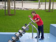 Ein Junge in einem roten Hemd dreht eine archimedische Schraube aus Metall in einem Außenbereich mit Gras und Bäumen.