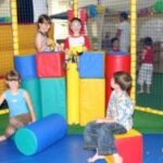 Vier Kinder spielen mit großen Schaumstoffblöcken in einem mit Netzen eingefassten Innenspielbereich. Andere Kinder sind im Hintergrund zu sehen.