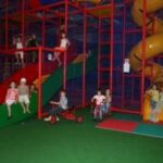 Kinder spielen auf Rutschen, Klettergerüsten und fahren auf einem Dreirad in einem Indoor-Spielplatz mit bunten Geräten und gepolsterten Böden.