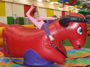 Ein Kind reitet auf einem großen roten aufblasbaren Stier in einem bunten Indoor-Spielbereich, hebt einen Arm und lächelt.