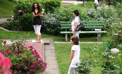 Drei Personen stehen in einem Gartenbereich in der Nähe einer Bank und von Blumenbeeten, die von Grün und rosa Blumen umgeben sind.