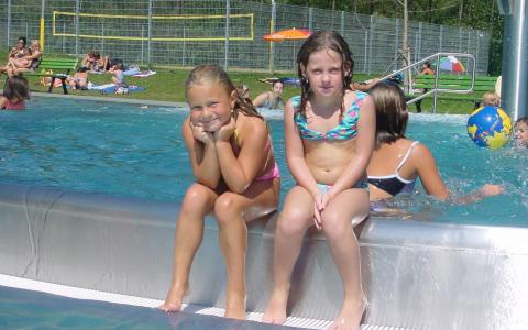 Zwei junge Mädchen in Badeanzügen sitzen an einem sonnigen Tag am Rande eines Freibads, während im Hintergrund andere Kinder schwimmen und spielen.