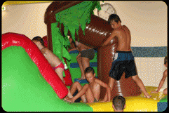 Mehrere Kinder in Badeanzügen spielen auf einer aufblasbaren Wasserrutsche in einer Halle.