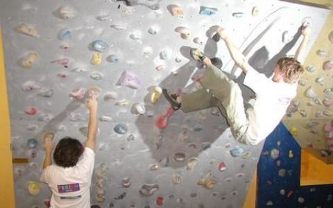 Zwei Personen klettern in der Halle an einer künstlichen Kletterwand, wobei eine Person höher klettert, während die andere nach den Griffen unten greift.