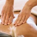 Person, die eine Rückenmassage erhält, wobei zwei Hände den nackten, teilweise mit einem Handtuch bedeckten oberen Rücken drücken und massieren.