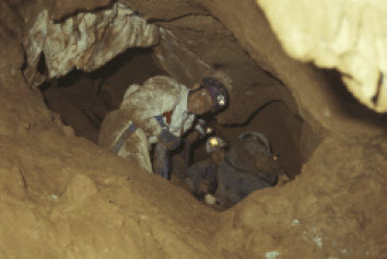 Drei Personen mit Helmen und Scheinwerfern erkunden einen engen, felsigen Höhlengang.