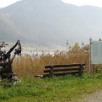 Eine Holzbank, eine Treibholzskulptur und ein Informationsschild stehen an einem See, im Hintergrund sind die Berge zu sehen.