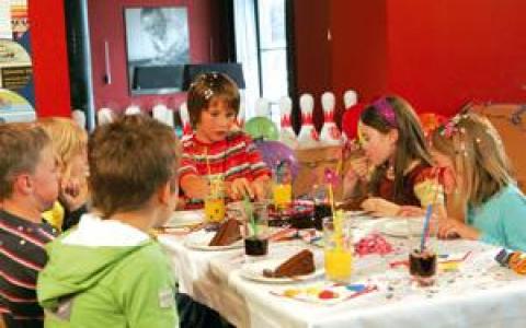 Kinder sitzen um einen Tisch herum, essen Kuchen und trinken Saft auf einer Geburtstagsfeier, im Hintergrund sind Partydekorationen und Bowlingkegel zu sehen.