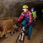 Menschen mit Helmen und Jacken fahren auf Fahrrädern in einem schwach beleuchteten Tunnel neben einem alten Bergbauwagen auf Schienen.
