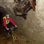 Zwei Radfahrer, die Helme und Sicherheitsausrüstung tragen, fahren mit ihren Fahrrädern durch einen schwach beleuchteten, felsigen Tunnel, über dem sich Bergbaumaschinen befinden.