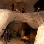 Mehrere Personen mit Stirnlampen erkunden eine felsige Höhle mit mehreren Fahrrädern, die an den Wänden und am Boden der Höhle lehnen.