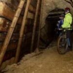 Zwei Personen mit Helmen und Sicherheitsausrüstung stehen mit ihren Fahrrädern in einem schwach beleuchteten, verstärkten Tunnel mit Holzstützen.