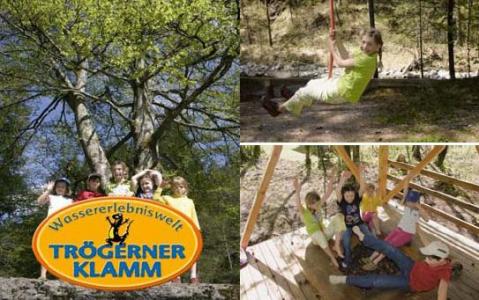 Kinder spielen in einem Naturpark, einige stehen hinter einem Schild mit der Aufschrift "Wassererlebniswelt Trögerner Klamm", andere freuen sich über Spielgeräte.