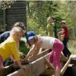 Mehrere Kinder spielen im Freien auf einer Holzkonstruktion in einem bewaldeten Gebiet, mit Bäumen und Grün im Hintergrund.