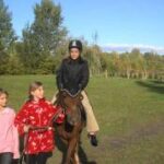 Eine Person, die einen Helm trägt, reitet auf einem Pferd, angeführt von zwei Kindern, auf einer Wiese im Freien mit Bäumen im Hintergrund.