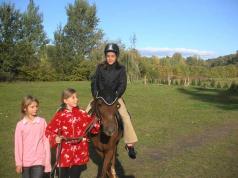 Eine Person, die einen Helm trägt, reitet auf einem Pferd, angeführt von zwei Kindern, auf einer Wiese im Freien mit Bäumen im Hintergrund.