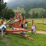 Kinder spielen auf einem hölzernen Spielplatz in einem grasbewachsenen Außenbereich mit Bäumen und Hügeln im Hintergrund.