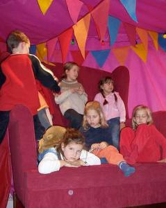 Sechs Kinder sitzen und lümmeln auf einem großen roten Stuhl in einem Zelt, das mit bunten dreieckigen Wimpeln geschmückt ist.