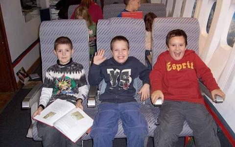 Drei Jungen sitzen zusammen in flugzeugähnlichen Sitzen; einer hält ein Buch, einer winkt und lächelt, ein anderer grinst breit. Hinter ihnen sitzen andere Kinder, die den Blick abwenden.