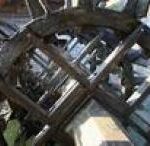 Nahaufnahme eines hölzernen Wasserrads, auf dessen Struktur das Sonnenlicht Schatten wirft und unter dem Wasser fließt.