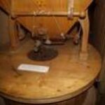 Nahaufnahme einer hölzernen Getreidemühle mit Trichter und Mahlwerk, die in einem Innenraum auf einem Holzsockel steht.