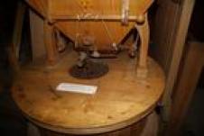 Nahaufnahme einer hölzernen Getreidemühle mit Trichter und Mahlwerk, die in einem Innenraum auf einem Holzsockel steht.