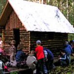 Eine Gruppe von Kindern und ein Erwachsener sitzen und stehen in der Nähe einer kleinen Holzhütte in einem Waldgebiet.