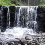 Ein kleiner Wasserfall fließt über eine Steinmauer in einen felsigen Bach, umgeben von Grün und Bäumen im Hintergrund.