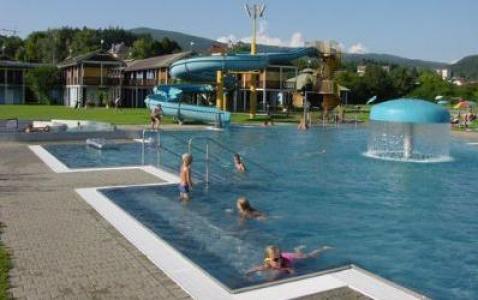 Kinder schwimmen in einem Freibad mit einer Wasserrutsche und einem pilzförmigen Springbrunnen. Bei klarem Himmel sind im Hintergrund Gebäude und Hügel zu sehen.
