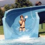 Ein Kind rutscht eine blaue Wasserrutsche hinunter und spritzt am Ende in ein Becken. Im Hintergrund sind Gras und Bäume zu sehen.