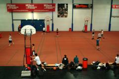 In einer Sporthalle wird auf mehreren Plätzen Basketball gespielt, wobei Bänke und Sportgeräte den Rand des Spielfelds säumen.