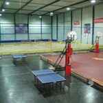 Hallensportanlage mit Basketballkörben, Tischtennisplatten und einem Sandvolleyballfeld. Bänke und Sportgeräte sind in dem Bereich zu sehen.