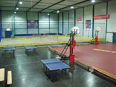 Hallensportanlage mit Basketballkörben, Tischtennisplatten und einem Sandvolleyballfeld. Bänke und Sportgeräte sind in dem Bereich zu sehen.