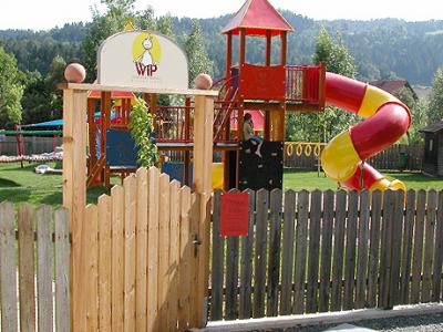 Ein eingezäunter Spielplatz mit einem "VIP"-Schild am Eingang, mit einer roten und gelben Spiralrutsche, Klettergerüsten und grünem Rasen.
