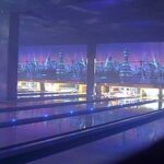 Eine Bowlingbahn mit leuchtenden blauen Lichtern, leeren Bahnen und futuristisch anmutender Wandmalerei im Hintergrund.