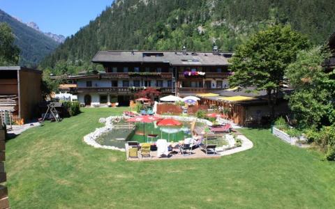 Ein grasbewachsener Hof mit Liegestühlen umgibt einen natürlichen Schwimmteich vor einem Gebäude im Lodge-Stil mit Balkonen, das von bewaldeten Bergen umgeben ist.
