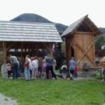 Eine Gruppe von Menschen, darunter Kinder und Erwachsene, versammelt sich in der Nähe von zwei Holzkonstruktionen in einem Außenbereich mit Gras und Bergen im Hintergrund.