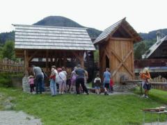 Eine Gruppe von Menschen, darunter Kinder und Erwachsene, versammelt sich in der Nähe von zwei Holzkonstruktionen in einem Außenbereich mit Gras und Bergen im Hintergrund.