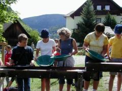 Fünf Personen stehen im Freien und wühlen mit grünen Pfannen nach Gold, während im Hintergrund Häuser und Berge zu sehen sind.